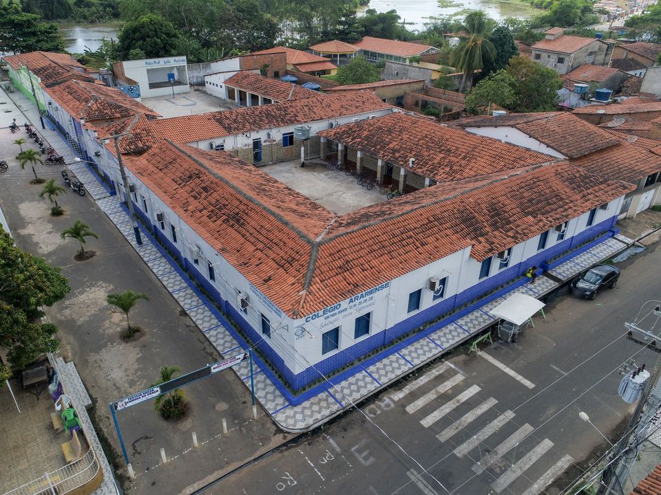 Vista aérea do Colégio Arariense, na Praça da Bandeira, em Arari: prédio em L com telhado de cerâmica, paredes brancas com faixa azul e o nome do colégio na esquina.
